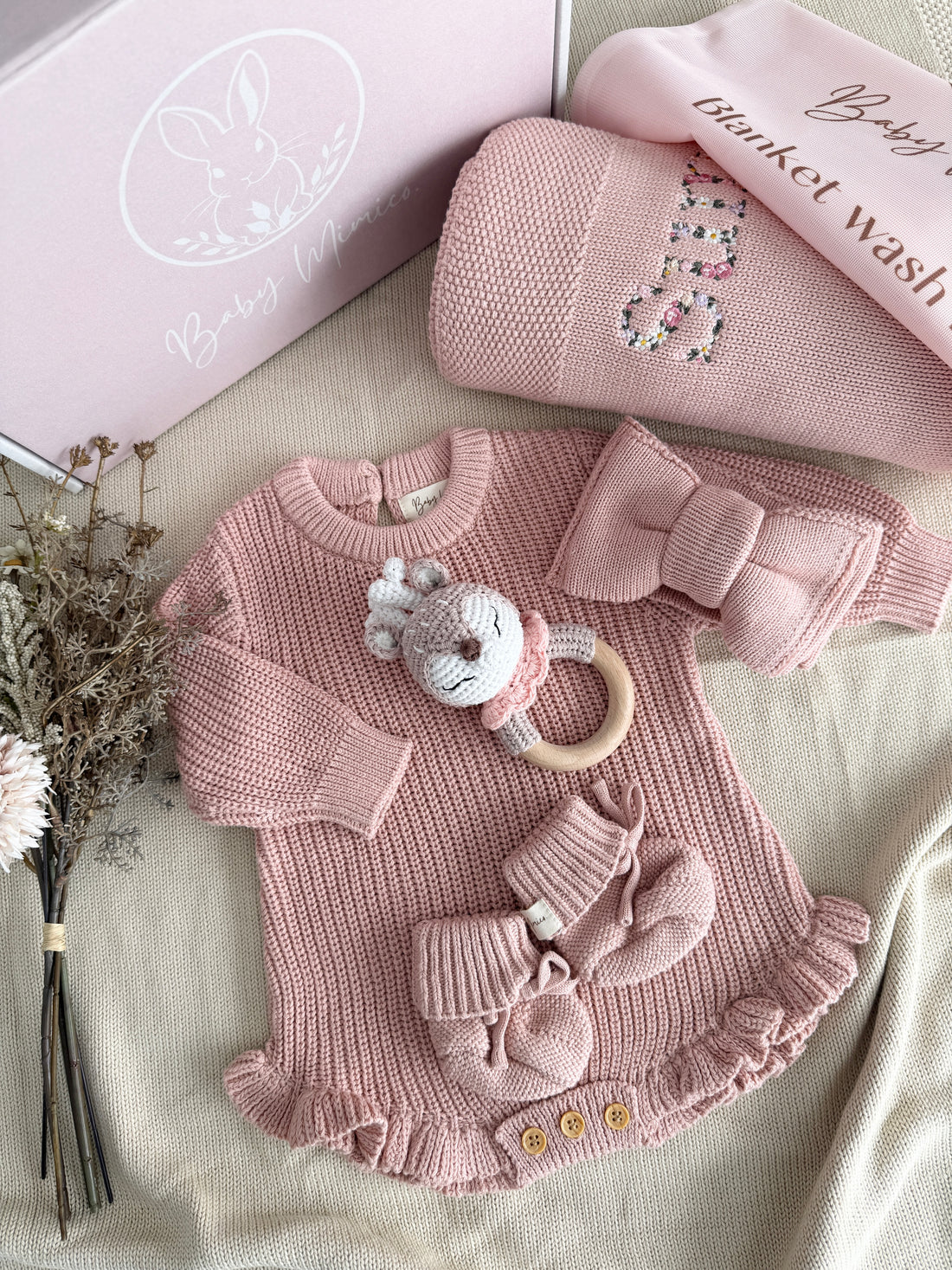 A coordinated baby knit set arranged on a beige fabric surface, including a pink long-sleeve romper with ruffle trim, matching booties, a bow headband, a plush rattle, and a folded pink blanket or wrap. A pink branded box and a folded pink item with &