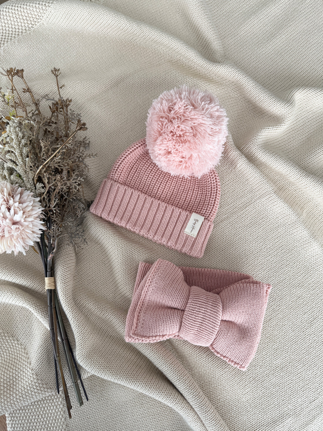 A baby knit hat and matching bow headband in dusty pink, styled on a beige textured blanket with dried flowers.