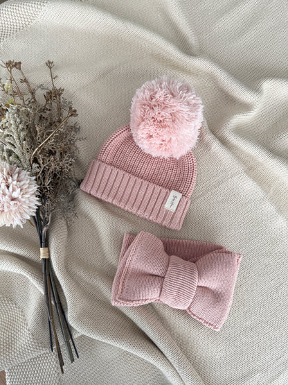 A baby knit hat and matching bow headband in dusty pink, styled on a beige textured blanket with dried flowers.