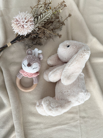 A soft plush bunny toy and a small crocheted animal rattle with a wooden ring, styled on a beige textured fabric with dried flowers.